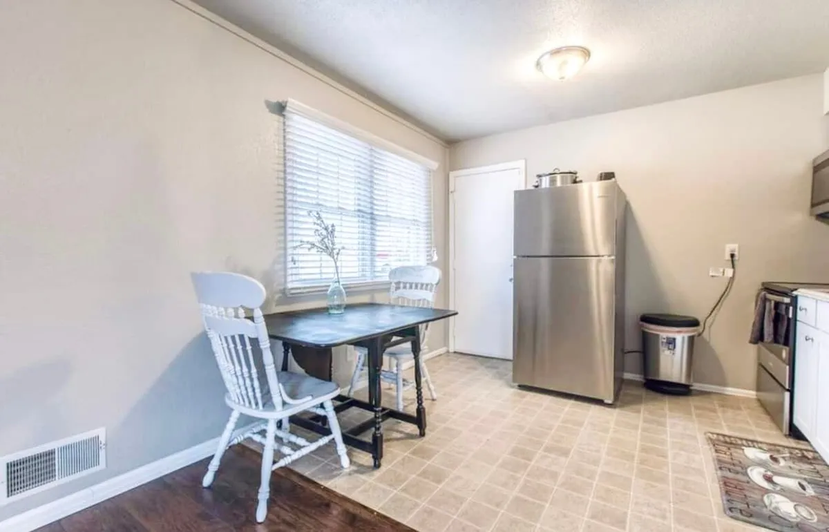 A cozy dining nook with a small dining table, two white chairs, and a stainless steel refrigerator in view. Sunlight pours in through a window with blinds, creating a bright and comfortable spot for shared meals or a quiet morning coffee.