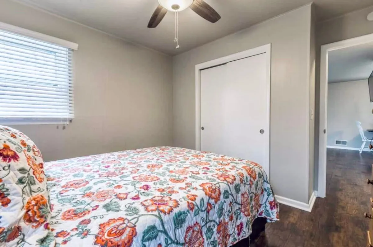 A peaceful bedroom scene showcasing a flower-patterned bedspread, a window with blinds letting in gentle daylight, and a closet door. The space is arranged to encourage rest and quiet moments, with a cozy reading nook by the window.