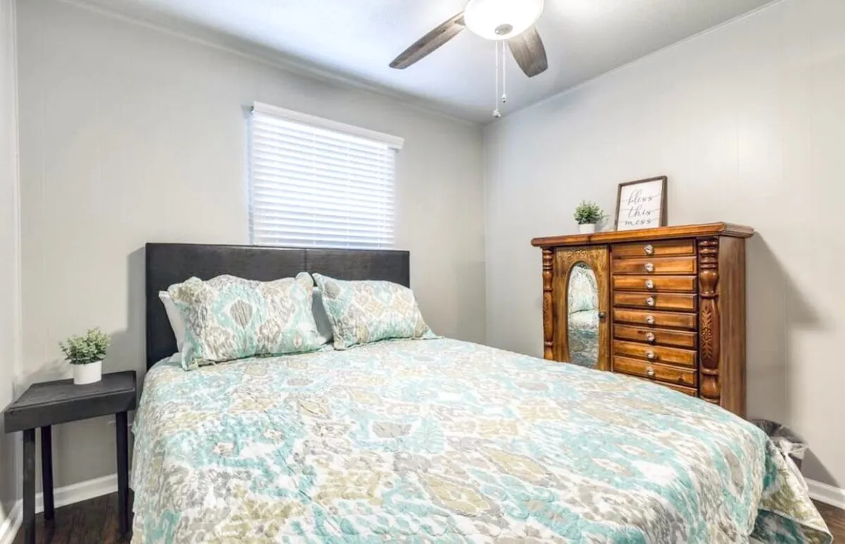 A tranquil bedroom scene featuring a bed with patterned bedding, a vintage dresser, and a small side table topped with a plant. Soft, peaceful morning light streams in, casting a cozy glow throughout the space. The overall mood is inviting and restful, perfect for winding down after a busy day.