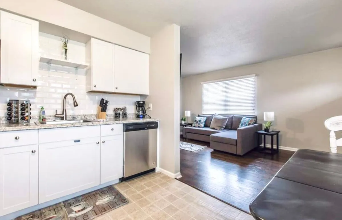 An open-concept view featuring a kitchen counter, dining table, sofa, and large windows. The kitchen seamlessly transitions into the dining area, highlighting the ease of moving meals from stove to table while also supporting entertaining and socializing.