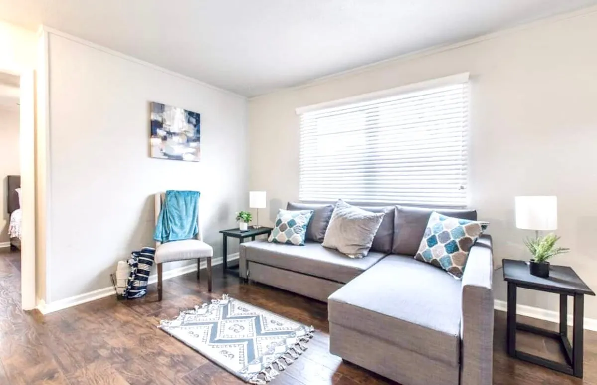 This image showcases a cozy living room with a plush sofa, decorative pillows, tasteful artwork on the wall, and inviting side tables atop a textured rug. The arrangement suggests a welcoming space for relaxing, reading, or engaging in casual conversation, with plenty of natural light filtering in.