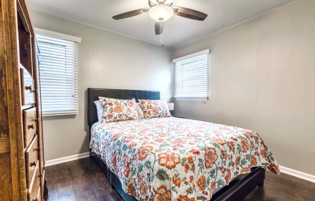 A warm, inviting bedroom with a bed dressed in floral bedding, a wooden dresser, and a window with blinds. Natural light floods the room, creating a soothing environment. The ceiling fan hints at comfort for all seasons, and the space feels both private and relaxing.